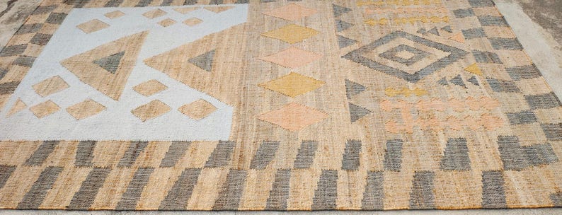 May include: A rectangular woven rug featuring a natural tan fiber base with geometric patterns in white, charcoal gray, mustard yellow, and soft peach. The design includes triangles, diamonds, and repeating block borders across the surface.