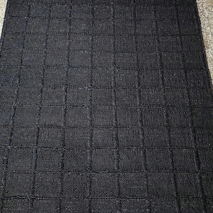 Puede incluir: Una alfombra rectangular negra con un dise&ntilde;o de cuadr&iacute;cula. La alfombra est&aacute; hecha de un material tejido, y la cuadr&iacute;cula est&aacute; formada por l&iacute;neas m&aacute;s oscuras que crean una apariencia texturizada. Adecuada para uso en interiores.