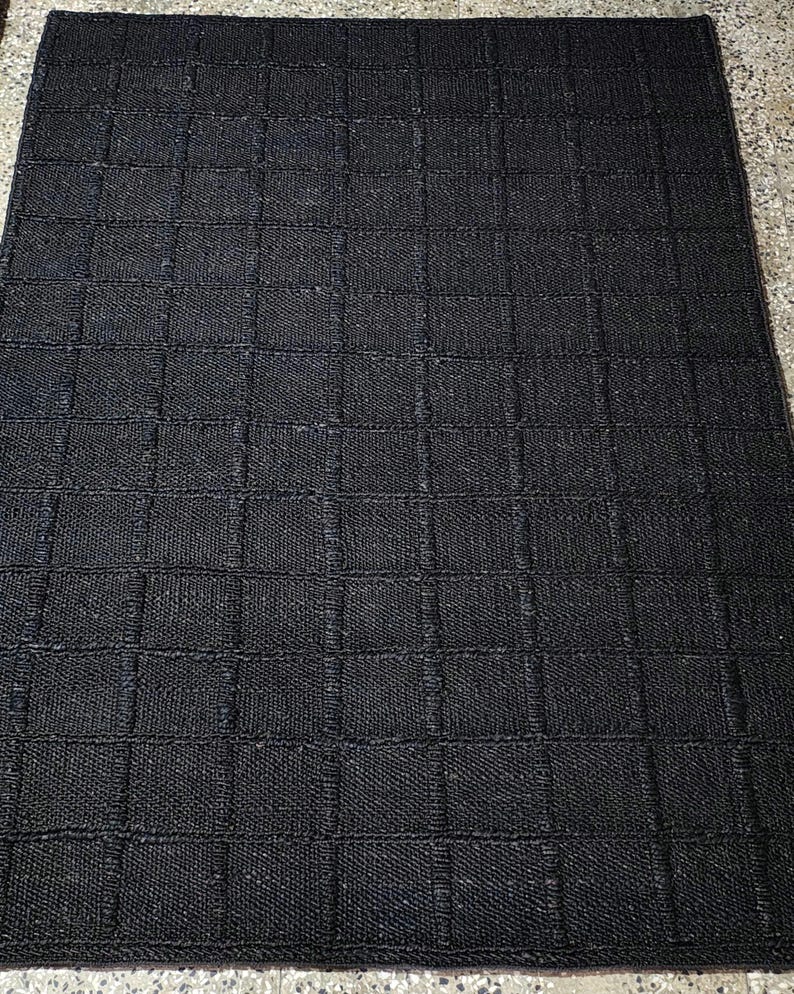 Puede incluir: Una alfombra rectangular negra con un dise&ntilde;o de cuadr&iacute;cula. La alfombra est&aacute; hecha de un material tejido, y la cuadr&iacute;cula est&aacute; formada por l&iacute;neas m&aacute;s oscuras que crean una apariencia texturizada. Adecuada para uso en interiores.