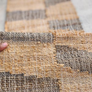 May include: A rectangular woven runner rug featuring a repeating geometric pattern of alternating tan and dark gray blocks. The rug is constructed from thick, textured natural fibers with a visible grid-like weave and finished edges.