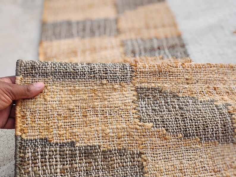 May include: A rectangular woven runner rug featuring a repeating geometric pattern of alternating tan and dark gray blocks. The rug is constructed from thick, textured natural fibers with a visible grid-like weave and finished edges.