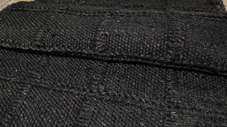 Puede incluir: Primer plano de una alfombra tejida de color gris oscuro con una superficie texturizada. La alfombra tiene un patr&oacute;n geom&eacute;trico con secciones elevadas y un brillo sutil. El material parece ser una fibra natural, posiblemente yute o similar.