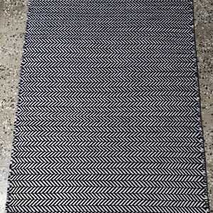May include: A rectangular area rug featuring a high-contrast black and white herringbone pattern. The woven textile displays a consistent zigzag chevron design across the entire surface, finished with short fringe edges on the top and bottom.