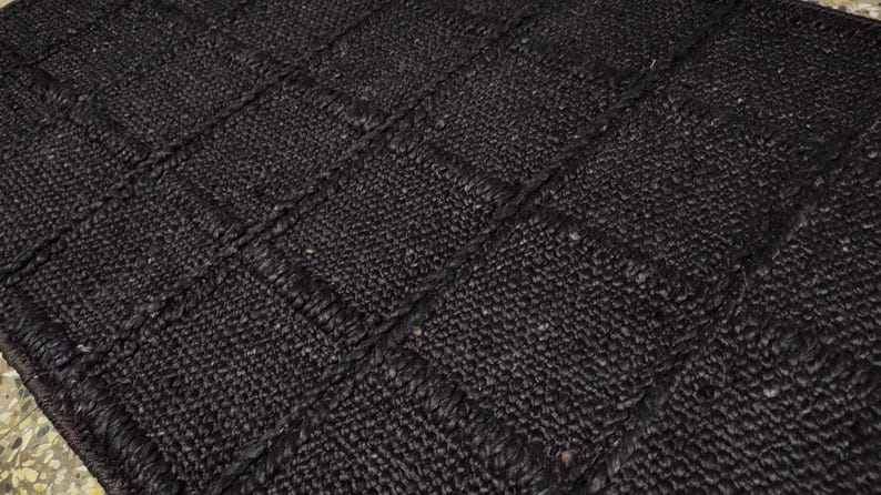 May include: A rectangular area rug featuring a dark black color and a woven texture. The rug is constructed with a grid pattern of square sections, each defined by thick, braided borders that create a raised, tactile surface across the entire piece.