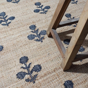 May include: A woven area rug with a natural tan base color featuring a repeating dark blue floral pattern. The rug has a textured, coarse surface and is partially covered by the wooden legs of a stool, showing the weave and print detail.