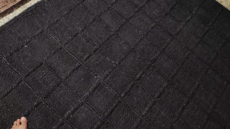 May include: A round, black area rug featuring a textured, woven surface with a prominent grid pattern formed by raised lines. The rug has a consistent dark charcoal tone and a dense, fibrous construction that creates a uniform, geometric appearance.