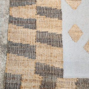 May include: A woven rug featuring a textured border of alternating tan and dark gray rectangular blocks. The central section is a light gray field decorated with a repeating pattern of tan diamond shapes, creating a distinct contrast.