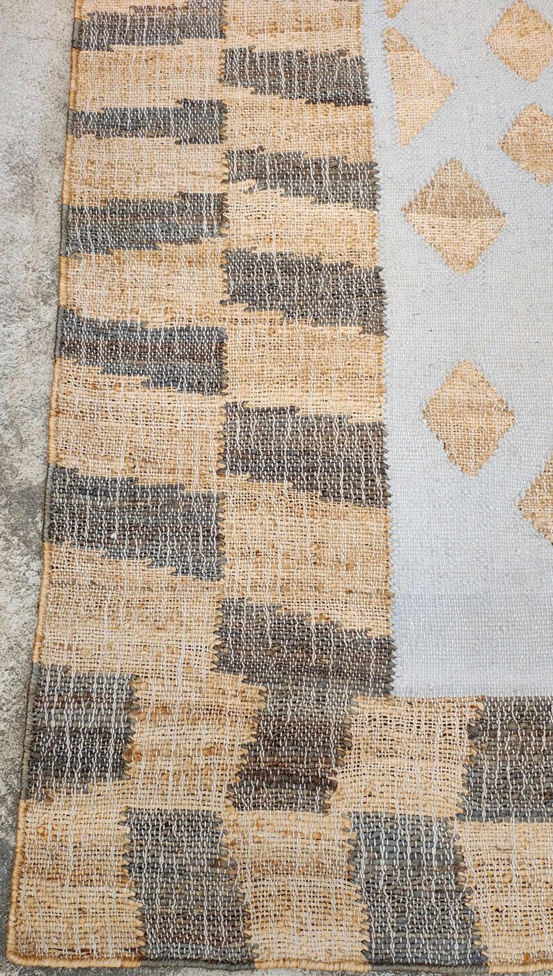 May include: A woven rug featuring a textured border of alternating tan and dark gray rectangular blocks. The central section is a light gray field decorated with a repeating pattern of tan diamond shapes, creating a distinct contrast.