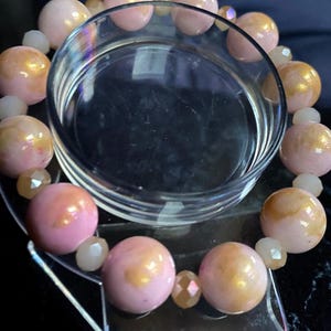 Könnte beinhalten: Ein rosafarbenes und goldenes Armband mit schillernden Perlen. Das Armband wird auf einem klaren Glasständer präsentiert.