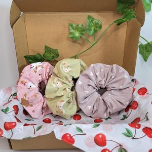 Peut inclure: Un ensemble de trois chouchous en tissu dans une boîte en carton. Un chouchou rose présente un motif de cerf, un autre est vert clair avec un motif similaire, et le troisième est mauve avec des cœurs blancs. La boîte est doublée de papier de soie imprimé de cerises.