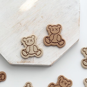 May include: Close-up of several small, tan teddy bear-shaped embellishments with stitched details and cross-shaped eyes. The bears are scattered on a white surface and a white, wooden, geometric-shaped board.