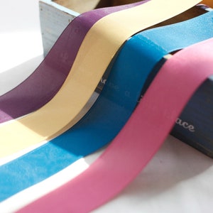 May include: Four rolls of satin ribbon in different colors: purple, yellow, blue, and pink. The ribbons are arranged in a fan shape, with the purple ribbon on top and the pink ribbon on the bottom.