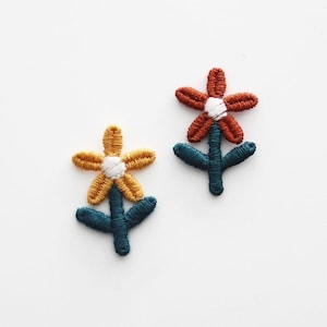 以下が含まれることがあります： 2つの刺繍された花のパッチ。1つの花はオレンジ色の花びら、白い中心、ティール色の茎があります。もう1つの花は黄色の花びら、白い中心、ティール色の茎があります。花は白い背景にあります。