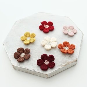 May include: A collection of embroidered flower appliques in various colors, including red, yellow, brown, and orange, with white and silver centers. The flowers are arranged on a white, hexagonal wooden surface. These decorative floral patches are ideal for crafting and embellishments.