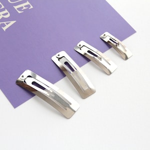 May include: Four silver metal hair clips of varying sizes. The clips are arranged in a row from largest to smallest on a white and purple surface.