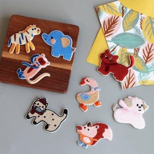 May include: A collection of embroidered animal patches including a zebra, blue elephant, monkey, red cat, goose, spotted cat with a fish, white dog with a pink bow, and a hedgehog. The patches are arranged on a wooden board and a leaf-patterned card.