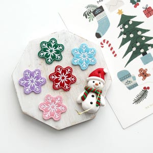 May include: A collection of colorful snowflake patches in green, blue, purple, red, and pink, arranged on a white wooden surface. A small snowman figurine with a red hat and green scarf is also present, along with Christmas-themed illustrations.