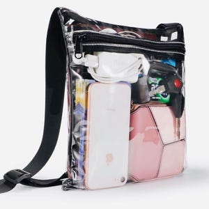 Peut inclure: Sac bandouli&egrave;re transparent avec une bandouli&egrave;re et une fermeture &eacute;clair noires. Le sac contient un smartphone blanc, un sac &agrave; main rose, un chargeur blanc et d'autres articles. Le sac est con&ccedil;u pour &ecirc;tre transparent, permettant une visibilit&eacute; facile du contenu.