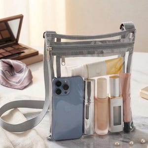 Peut inclure: Un sac bandouli&egrave;re transparent avec une bandouli&egrave;re et une fermeture &eacute;clair grises, rempli d'un smartphone bleu, de maquillage et de produits de soin. Le sac est pos&eacute; sur une surface en marbre, avec une palette de maquillage et une &eacute;charpe en arri&egrave;re-plan.