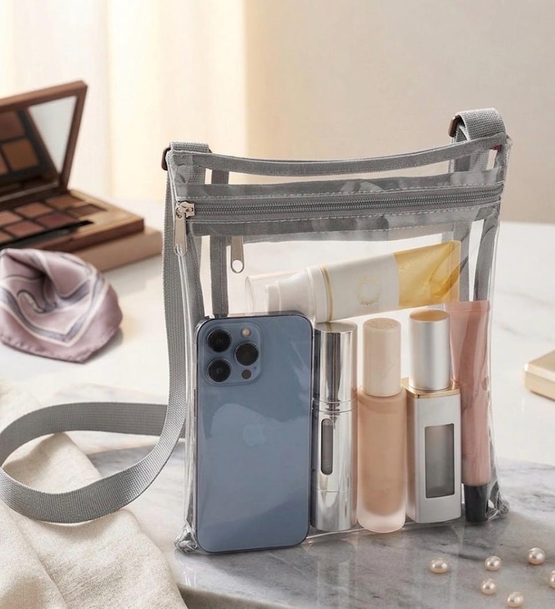 Peut inclure: Un sac bandouli&egrave;re transparent avec une bandouli&egrave;re et une fermeture &eacute;clair grises, rempli d'un smartphone bleu, de maquillage et de produits de soin. Le sac est pos&eacute; sur une surface en marbre, avec une palette de maquillage et une &eacute;charpe en arri&egrave;re-plan.