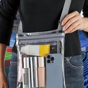 Peut inclure: Sac bandouli&egrave;re transparent avec une sangle grise, contenant un t&eacute;l&eacute;phone, des cosm&eacute;tiques et un tube de cr&egrave;me solaire. Le sac est clair, permettant de voir le contenu. Le texte en haut indique: TRANSPARENT BODY TO GET THROUGH SECURITY.