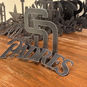 May include: Metal cutouts of the San Diego Padres logo and the word "Padres" in a stylized font. The cutouts are dark gray and sit on a wooden surface. Other metal cutouts are in the background.
