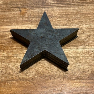 以下が含まれることがあります： 暗い灰色の仕上げを施した、素朴な金属製の星が5つ積み重ねられています。星は木製の表面に、先端を上に向けて配置されています。