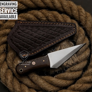 May include: A small, handcrafted knife with a polished steel blade and a dark wooden handle. The knife rests on a thick, coiled rope next to a brown leather sheath with a diamond pattern. Text in the image reads "ENGRAVING SERVICE AVAILABLE."