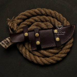 May include: A handcrafted hunting knife with a bone handle and a dark brown leather sheath secured with brass studs. The knife rests on a coiled rope, showcasing its rugged design. The knife is a tool for outdoor activities.