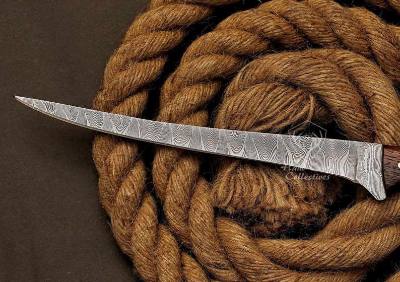 May include: A curved, silver-colored knife with a wooden handle and a patterned blade. The knife is resting on a brown rope.