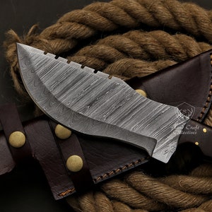 May include: A Damascus steel knife with a curved blade and serrated edge, resting on a brown leather sheath with brass rivets. The knife has a dark wooden handle and a unique pattern on the blade. A thick rope is in the background.
