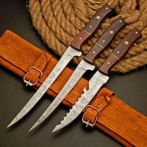 May include: Set of three Damascus steel knives with wooden handles and brass rivets. The blades have a unique patterned design. The knives are displayed with a brown leather sheath and a rope background.