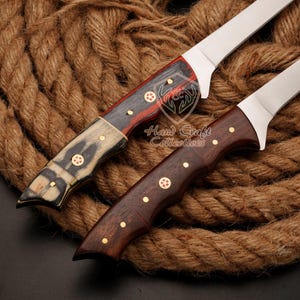 May include: Two knives with wooden handles and stainless steel blades. The handles are made of different types of wood, one with a red and black pattern and the other with a dark brown wood. The knives are resting on a rope background. "Hand Craft Collectives" is printed on the background.