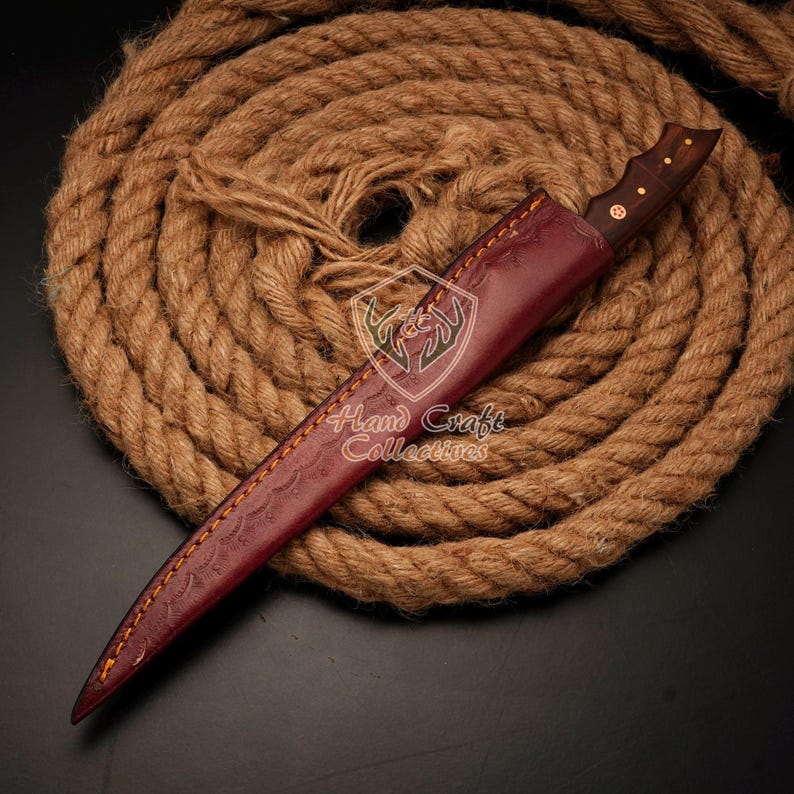 May include: A brown leather sheath with a decorative pattern and a wooden handle knife. The knife is wrapped in a brown leather sheath with a decorative pattern. The sheath is lying on a coiled rope.