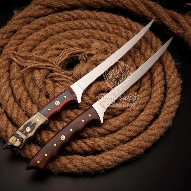 May include: Two long, thin, silver knives with wooden handles. The knife on the left has a black and red handle with brass accents. The knife on the right has a dark brown handle with brass accents. The knives are lying on a brown rope.