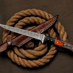 May include: A Damascus steel hunting knife with a brown leather sheath and a wooden handle with black and red accents. The knife is resting on a coil of brown rope.