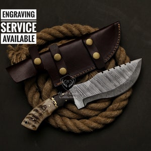 May include: A Damascus steel knife with a brown leather sheath. The knife has a patterned blade and a bone handle. The sheath is secured with brass-colored studs. Text on the image reads "ENGRAVING SERVICE AVAILABLE."