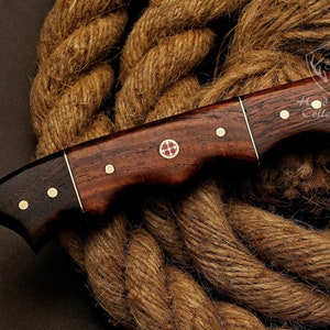 May include: A brown wooden handled knife with a Damascus steel blade. The handle has brass accents and a red cross design. The knife is resting on a brown rope.