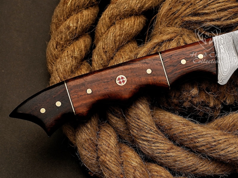 May include: A brown wooden handled knife with a Damascus steel blade. The handle has brass accents and a red cross design. The knife is resting on a brown rope.