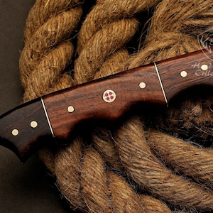 May include: A brown wooden handled knife with a Damascus steel blade. The handle has brass accents and a red cross design. The knife is resting on a brown rope.