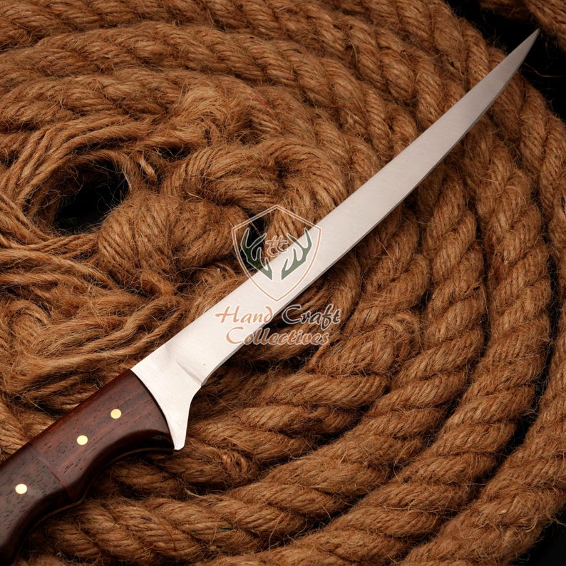 May include: A long, thin, silver knife with a wooden handle. The handle has a dark brown color and is decorated with brass accents. The knife is lying on a brown rope background.
