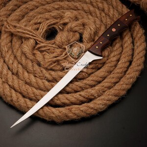 May include: A long, thin, silver knife with a wooden handle. The handle has a dark brown wood with a red cross design. The knife is lying on a brown rope.