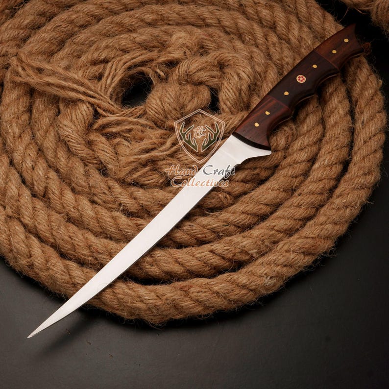 May include: A long, thin, silver knife with a wooden handle. The handle has a dark brown wood with a red cross design. The knife is lying on a brown rope.