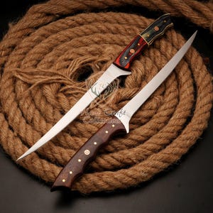 May include: Two stainless steel fishing knives with wooden handles. The handles are decorated with red, black, and gold accents. The knives are lying on a brown rope background.