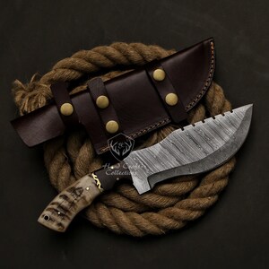 May include: A handcrafted hunting knife with a patterned steel blade and a bone handle. The knife is paired with a brown leather sheath secured with brass rivets. The knife and sheath rest on a coil of rope.