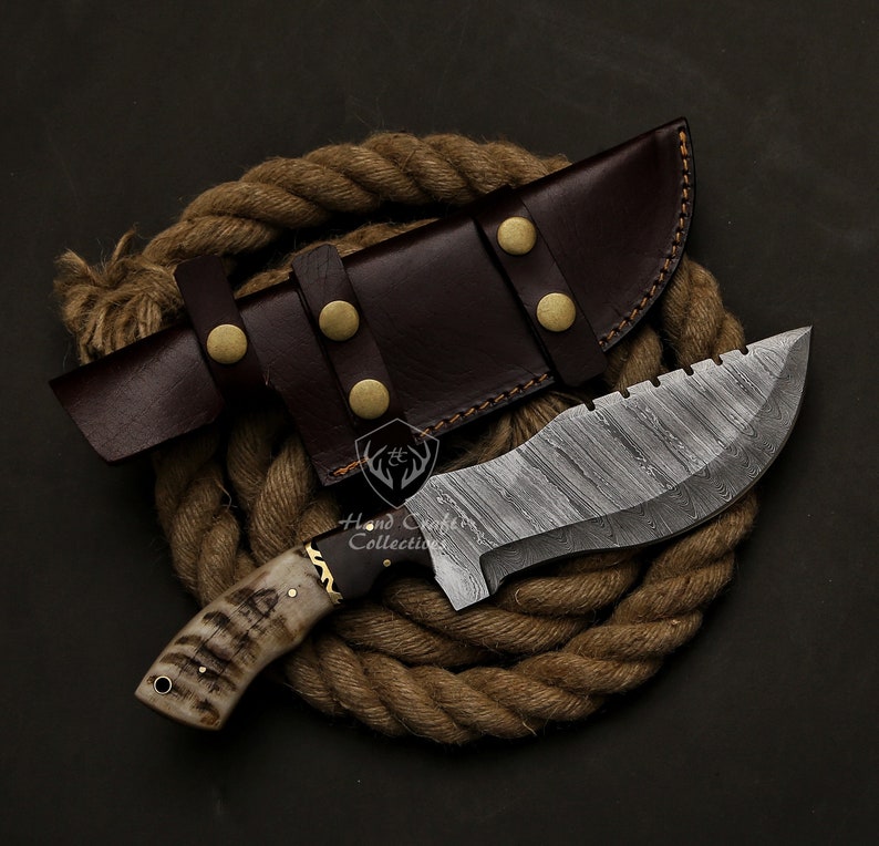 May include: A handcrafted hunting knife with a patterned steel blade and a bone handle. The knife is paired with a brown leather sheath secured with brass rivets. The knife and sheath rest on a coil of rope.