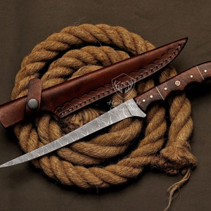 May include: A Damascus steel fishing knife with a brown wooden handle and a leather sheath. The knife is lying on a coil of brown rope.