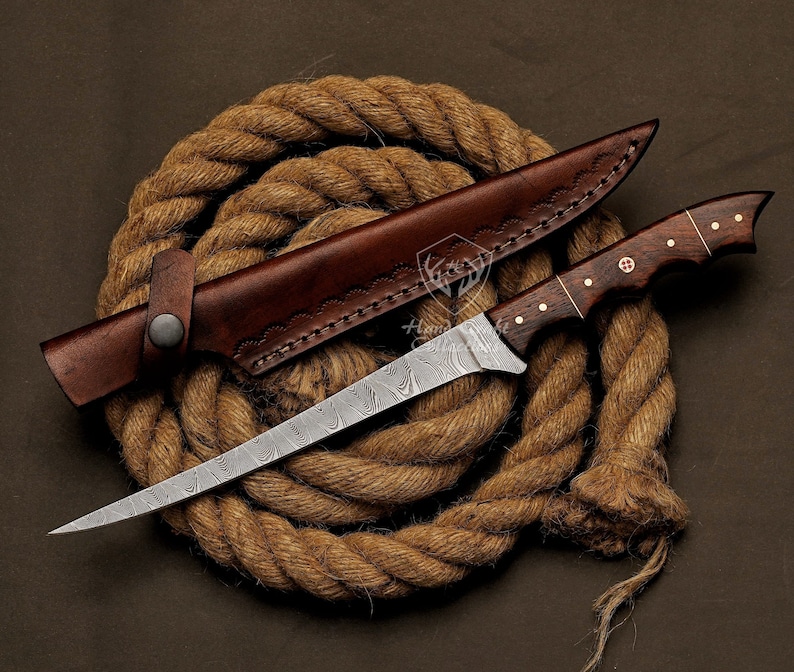 May include: A Damascus steel fishing knife with a brown wooden handle and a leather sheath. The knife is lying on a coil of brown rope.