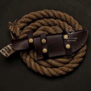 May include: A handcrafted hunting knife with a bone handle and a dark brown leather sheath secured with brass studs. The knife is resting on a coiled rope, showcasing a rustic aesthetic. The knife is a great gift for outdoor enthusiasts.