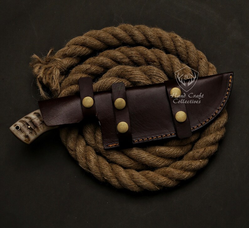 May include: A handcrafted hunting knife with a bone handle and a dark brown leather sheath secured with brass studs. The knife is resting on a coiled rope, showcasing a rustic aesthetic. The knife is a great gift for outdoor enthusiasts.
